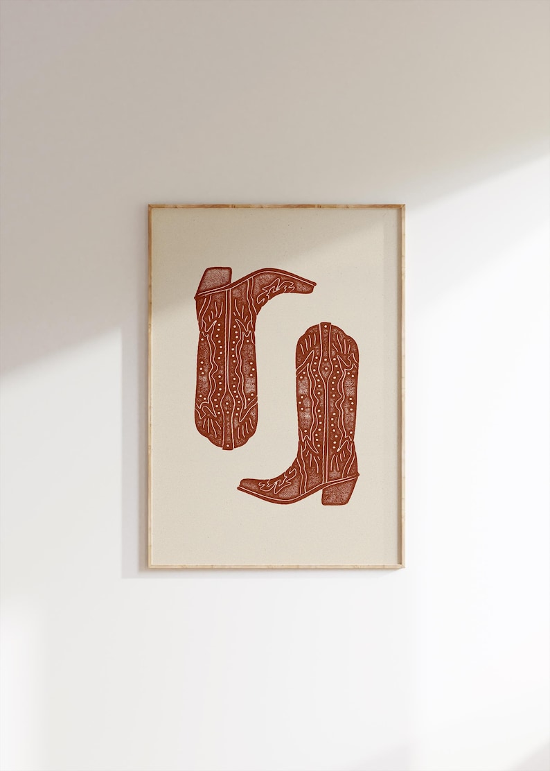Pu&ograve; includere: Una stampa incorniciata di due stivali da cowboy marroni con disegni intricati. Gli stivali sono su uno sfondo crema.