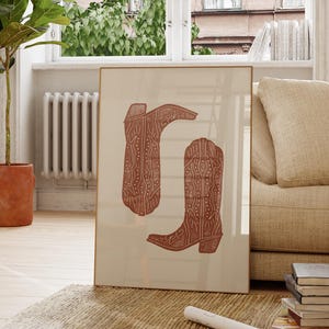 Pu&ograve; includere: Una stampa incorniciata di due stivali da cowboy marroni con motivi intricati. Gli stivali sono su uno sfondo crema.