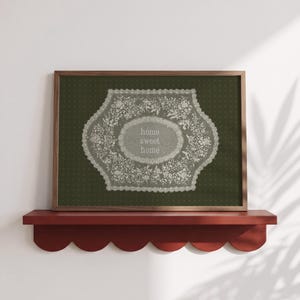 May include: A rectangular framed print featuring a dark olive green background with a subtle dot pattern. Centered is an intricate white lace-like floral design surrounding the text "home sweet home" in a soft, light-coloured, serif typeface.