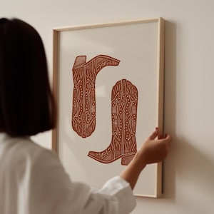 Pu&ograve; includere: Una stampa incorniciata di due stivali da cowboy marroni con motivi intricati. Gli stivali sono su uno sfondo bianco.