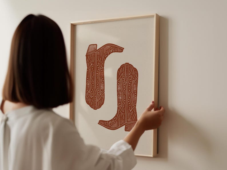 Pu&ograve; includere: Una stampa incorniciata di due stivali da cowboy marroni con motivi intricati. Gli stivali sono su uno sfondo bianco.