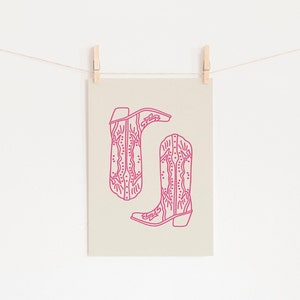 May include: A printable art print featuring a line drawing of two pink cowboy boots with intricate designs. The boots are on a beige background.