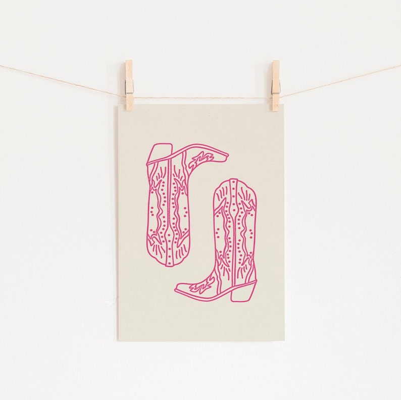 May include: A printable art print featuring a line drawing of two pink cowboy boots with intricate designs. The boots are on a beige background.