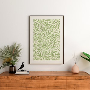 May include: A framed print featuring a green and white floral design hangs above a wooden dresser. The dresser has a vase with a green plant and a black bird figurine on top.