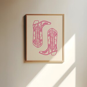 May include: A framed print of two pink cowboy boots with intricate designs. The boots are outlined in a darker pink and are set against a white background.