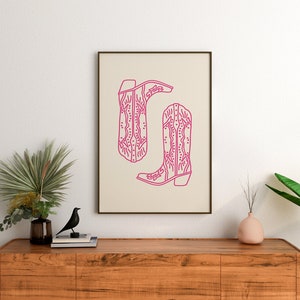 May include: A framed print of a line drawing of two pink cowboy boots with intricate designs. The boots are on a beige background.
