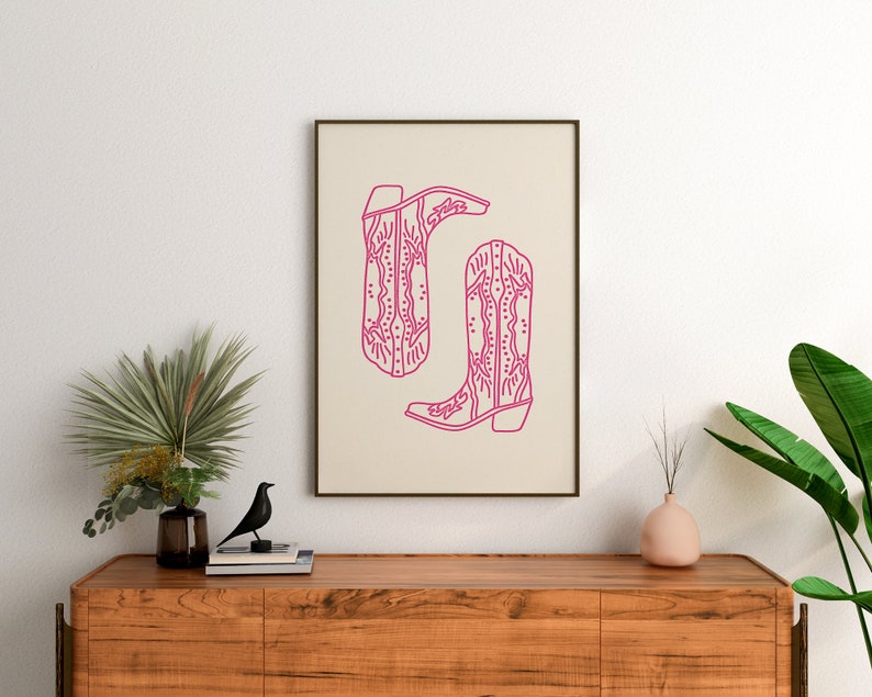 May include: A framed print of a line drawing of two pink cowboy boots with intricate designs. The boots are on a beige background.