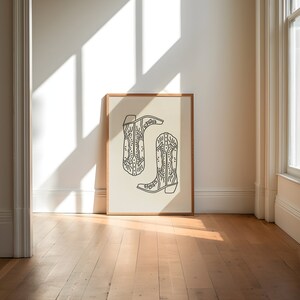 May include: A framed print of a line drawing of two cowboy boots with intricate designs. The boots are on a cream background.