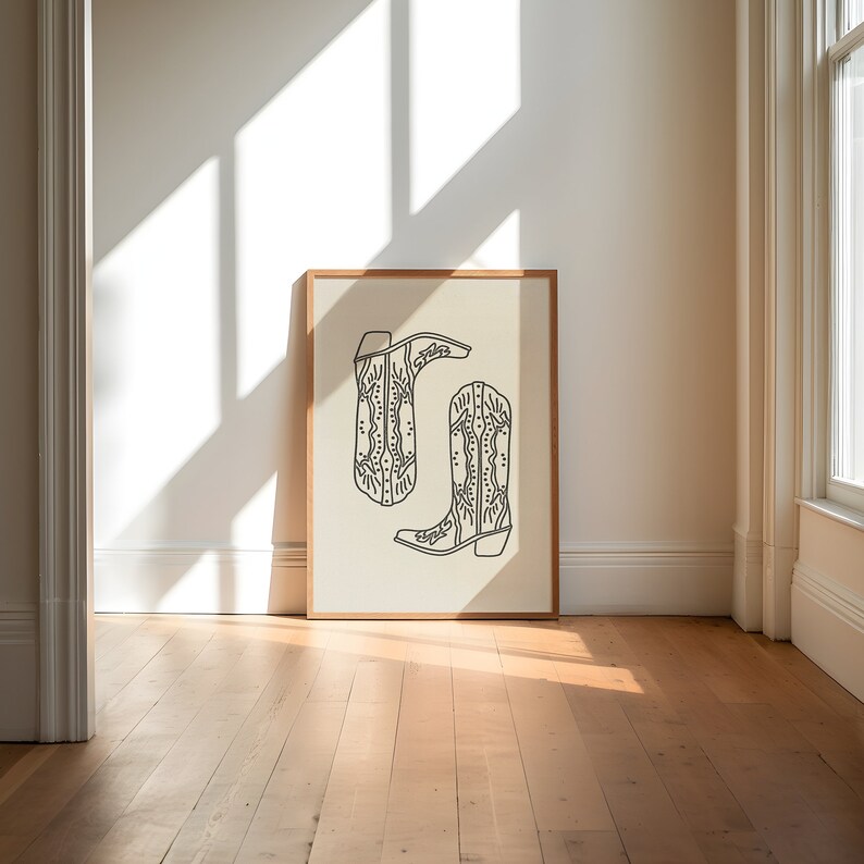 May include: A framed print of a line drawing of two cowboy boots with intricate designs. The boots are on a cream background.