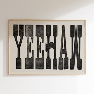 Peut inclure: Une affiche encadrée avec le mot "YEEHAW" en grosses lettres noires. Les lettres ont un aspect texturé et vieilli sur un fond beige clair. Le cadre est de couleur bois clair.