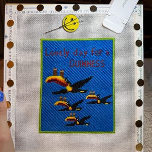 May include: Needlepoint canvas with a blue background and the text "Lonely day for a Guinness." Features a design of toucans in flight. The canvas is framed with a white border and gold-colored tacks.