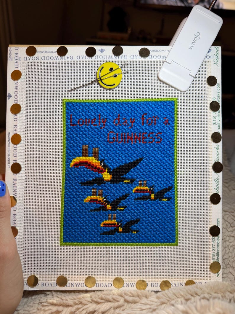 May include: Needlepoint canvas with a blue background and the text "Lonely day for a Guinness." Features a design of toucans in flight. The canvas is framed with a white border and gold-colored tacks.