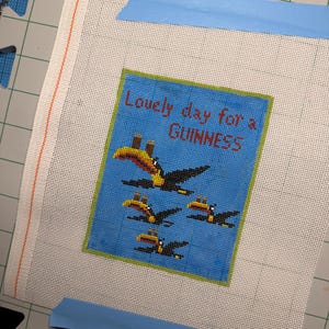 May include: Needlepoint canvas with a blue background and the text "Lovely day for a Guinness." Features a design of toucans. The canvas is taped to a green cutting mat with blue tape.
