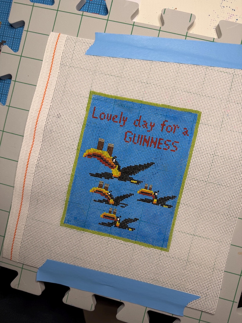 May include: Needlepoint canvas with a blue background and the text "Lovely day for a Guinness." Features a design of toucans. The canvas is taped to a green cutting mat with blue tape.