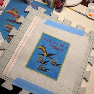 May include: A needlepoint canvas with a blue background featuring toucans and the text "Lovely day for a GUINNESS." The canvas is taped to a gray puzzle mat, surrounded by paint bottles and art supplies.