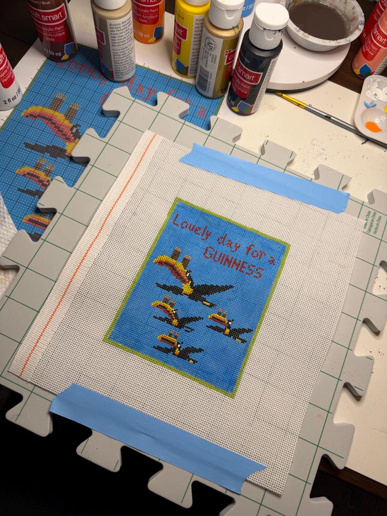 May include: A needlepoint canvas with a blue background featuring toucans and the text "Lovely day for a GUINNESS." The canvas is taped to a gray puzzle mat, surrounded by paint bottles and art supplies.