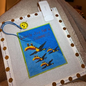 May include: A cross-stitch canvas with a blue background and toucan design, with the text "Lovely day for a Guinness". The canvas is secured with brown tacks and a blue thread. A white clip-on light is attached.