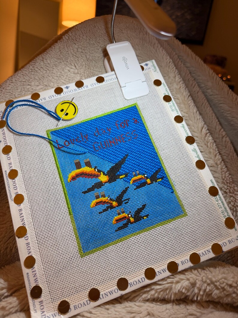 May include: A cross-stitch canvas with a blue background and toucan design, with the text "Lovely day for a Guinness". The canvas is secured with brown tacks and a blue thread. A white clip-on light is attached.