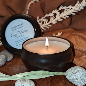 May include: A black candle jar with a lit candle inside. The candle jar is labeled "Hardy Wicks Candle Company Easy Whiskey 6.5 oz (187 g)". The jar is sitting on a wooden surface with a few small rocks and a green leaf.