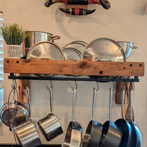 Könnte beinhalten: Ein rustikales Holzregal mit einem schwarzen Metall-Topfhalter, der darunter hängt. Das Regal ist mit verschiedenen Größen von Edelstahltöpfen und -pfannen gefüllt. Ein Paar rote und schwarze Kuhhörner ist an der Wand über dem Regal angebracht.