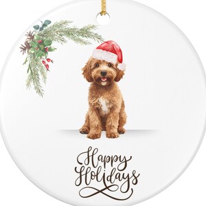 Könnte beinhalten: Runder, weißer Weihnachtsschmuck mit goldenem Band. Es zeigt einen braunen Hund mit Weihnachtsmütze. Die Worte "Happy Holidays" sind in einer Schrift unter dem Hund geschrieben. Grünzeug mit roten Beeren befindet sich auf der linken Seite.