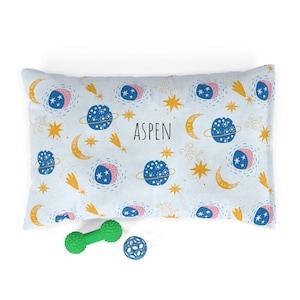 May include: A rectangular pet bed with a celestial pattern featuring planets, stars, and moons in blue, yellow, and pink on a white background. The name "ASPEN" is printed in black. Two green pet toys are in the foreground.
