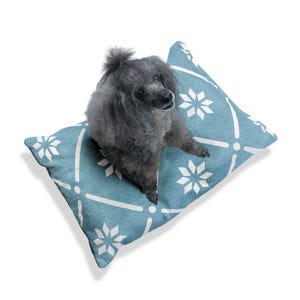 May include: A small gray dog sitting on a blue and white patterned dog bed. The bed has a white snowflake design.