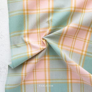 May include: A woven fabric featuring a plaid pattern in soft pastel shades of mint green, pale pink, and mustard yellow, accented by thin white intersecting lines. The textile is draped to show folds and includes the text FABLEISM printed at the bottom.
