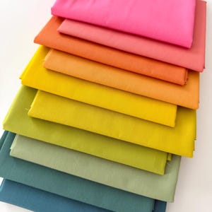 May include: A stack of folded fabric in a rainbow of colors, from hot pink to deep teal. The fabrics are arranged in a gradient, showcasing a variety of shades including orange, yellow, and green. The fabric appears to be cotton or a similar material.