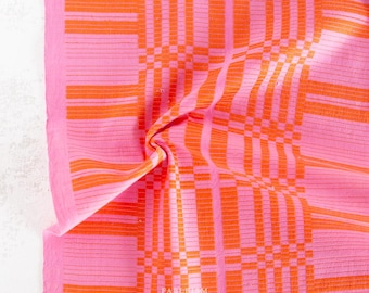 May include: Pink and orange patterned textile with a woven design. The fabric features a grid-like pattern of orange vertical and horizontal lines against a pink background. The textile is folded, creating shadows and texture.