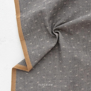 May include: A folded gray fabric with a repeating white cross pattern. The edge of the fabric has a tan border. The word "FABLEISM" is printed in white on the fabric.