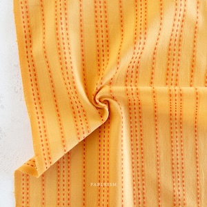 May include: Close-up of a folded yellow fabric with vertical orange stripes. The stripes are made of small, evenly spaced dashes. The fabric has a soft texture and a slight sheen. The word "FABLEISM" is printed in the bottom right corner.