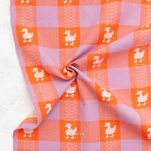 May include: A woven fabric featuring a checkered pattern in bright orange and light purple. The textile is decorated with a repeating white pixelated duck motif arranged in vertical columns, interspersed with thin, vertical zigzag line patterns.