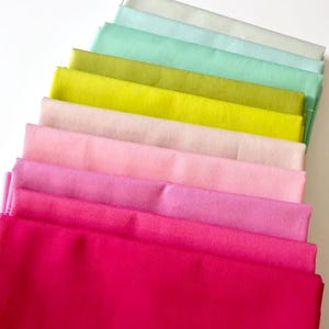 May include: A collection of folded fabric in a spectrum of colors, ranging from light pastels to vibrant hues. The arrangement showcases shades of pink, green, and yellow, creating a visually appealing display of textiles. The fabrics are neatly stacked, highlighting their smooth texture.