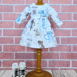 May include: A white doll dress with a cat print in blue, gray, and brown. The dress has long sleeves and a flared skirt. White doll boots with black laces are at the base of the wooden stand.