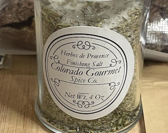May include: A clear glass jar with a lid, filled with dried herbs. A white label with black text reads "Herbes de Provence Finishing Salt, Colorado Gourmet Spice Co." The net weight is 4 oz. The jar sits on a wooden surface.