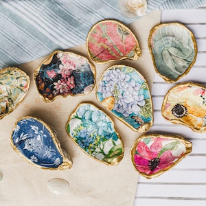 May include: A collection of decorative oyster shell dishes with gold-painted edges. Each shell features a unique floral design in various colors, including pink, blue, and yellow. The shells are arranged on a neutral-toned surface.