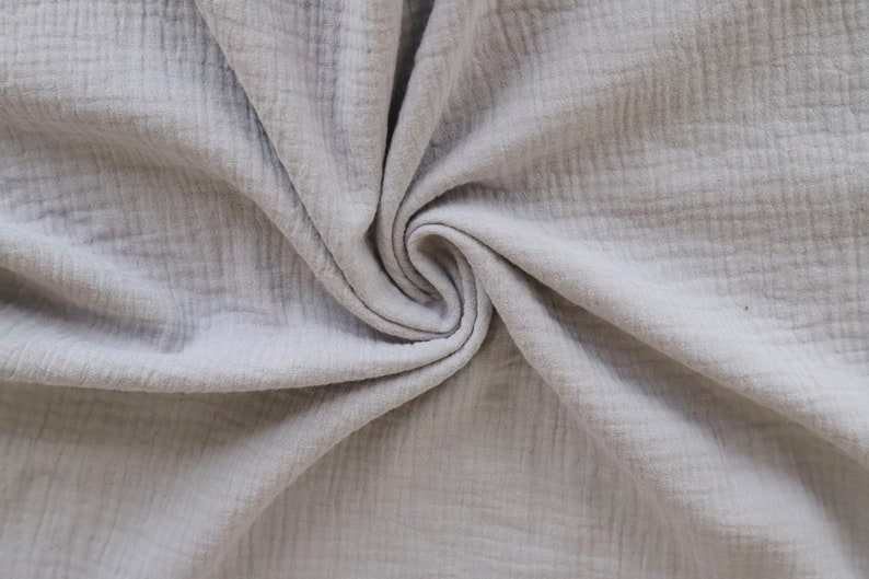 May include: A close-up of a soft, light gray fabric with a crinkled texture. The fabric is folded and draped, creating a swirling pattern.