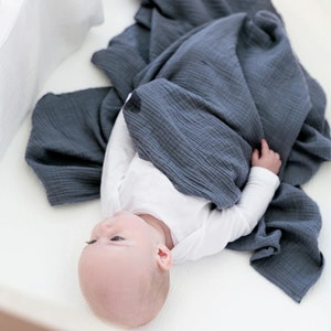 May include: A baby is lying in a crib wrapped in a dark blue muslin swaddle blanket. The blanket is made of a soft, breathable fabric. The baby is wearing a white onesie. The text "Fabric: #14 Indigo" is visible in the bottom right corner of the image.