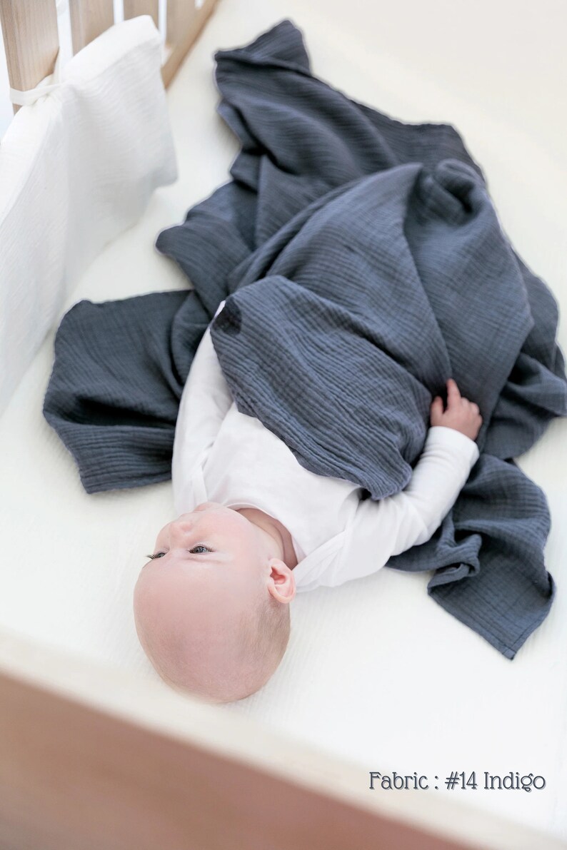 May include: A baby is lying in a crib wrapped in a dark blue muslin swaddle blanket. The blanket is made of a soft, breathable fabric. The baby is wearing a white onesie. The text "Fabric: #14 Indigo" is visible in the bottom right corner of the image.