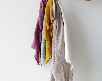 May include: A hand holding a stack of five different coloured muslin cloths. The cloths are in shades of pink, blue, yellow, beige, and white.
