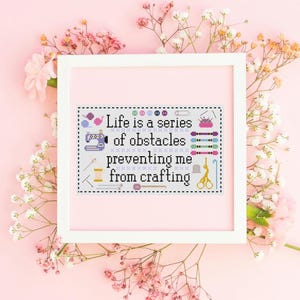 May include: A framed cross-stitch piece with the text "Life is a series of obstacles preventing me from crafting." The design includes sewing tools and a sewing machine. The frame is white, and the background is pink with floral accents.