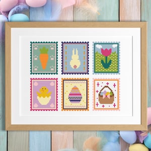 May include: A framed artwork featuring six cross-stitch designs with Easter themes. The designs include a carrot, bunny, tulip, chick, decorated egg, and a basket of eggs. The artwork is framed in a light brown wooden frame.