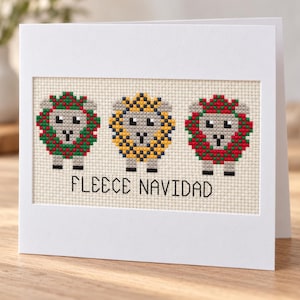 May include: A white greeting card featuring a cross-stitch design of three sheep on a cream-colored fabric background. The sheep are decorated in red, green, and yellow patterns. Below the sheep, the text reads FLEECE NAVIDAD in black letters.
