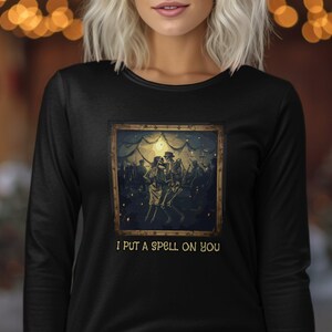 May include: Black long-sleeve shirt featuring a framed image of skeletons dancing. The image has a vintage aesthetic with a dark color palette. The text "I PUT A SPELL ON YOU" is printed below the image.