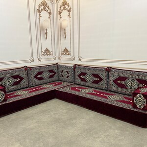 May include: A burgundy velvet L-shaped sofa with intricate patterned cushions. The sofa is set against a white wall with ornate gold trim.