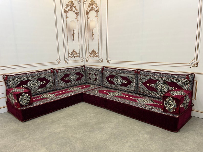 May include: A burgundy velvet L-shaped sofa with intricate patterned cushions. The sofa is set against a white wall with ornate gold trim.