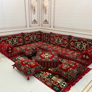 May include: A red and green patterned floor seating set with multiple cushions and pillows. The set includes a large L-shaped sofa with multiple cushions and pillows, as well as several smaller square cushions. The set is arranged on a white floor.