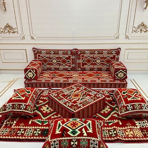 Puede incluir: Un conjunto de cojines de suelo con estampado rojo y verde con un puf a juego. El conjunto incluye un cojín de sofá grande, cuatro cojines más pequeños y un puf cuadrado. Los cojines y el puf están cubiertos de una tela roja con un patrón geométrico en verde, blanco y negro.