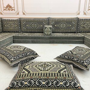May include: A gray and black patterned floor cushion set with a traditional Moroccan design. The set includes a large L-shaped sofa, four square cushions, and a small square ottoman.